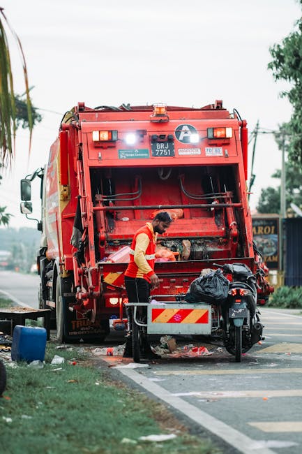 A middle-aged woman with short dark hair, dressed in casual dark clothing, is seen leaning over to dispose of a large white plastic bag filled with waste into a shiny, cylindrical stainless steel rubb